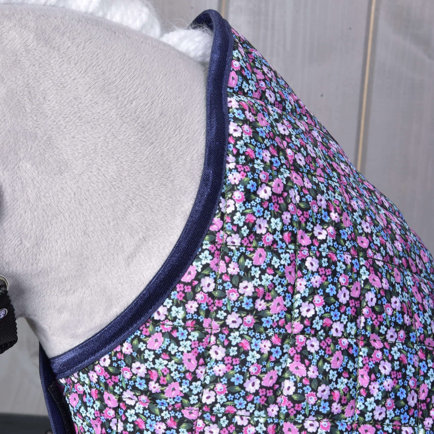 Detail koňa s prešívanou dekou Hobby Horse LarDen Blanket Flower s tmavomodrým lemom a malými ružovými, fialovými, bielymi a modrými kvetmi na tmavom podklade. Pozadie tvorí svetlé drevo.