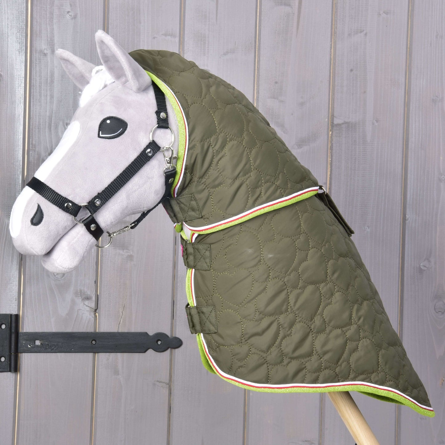 Sivý Hobby Horse s čiernou uzdečkou má na sebe Deku s odopínateľným krkom Rubby od Hobby Horse LarDen, zelenú kvalitnú deku so srdiečkovým vzorom a farebným lemom, zobrazenú pred drevenou stenou.