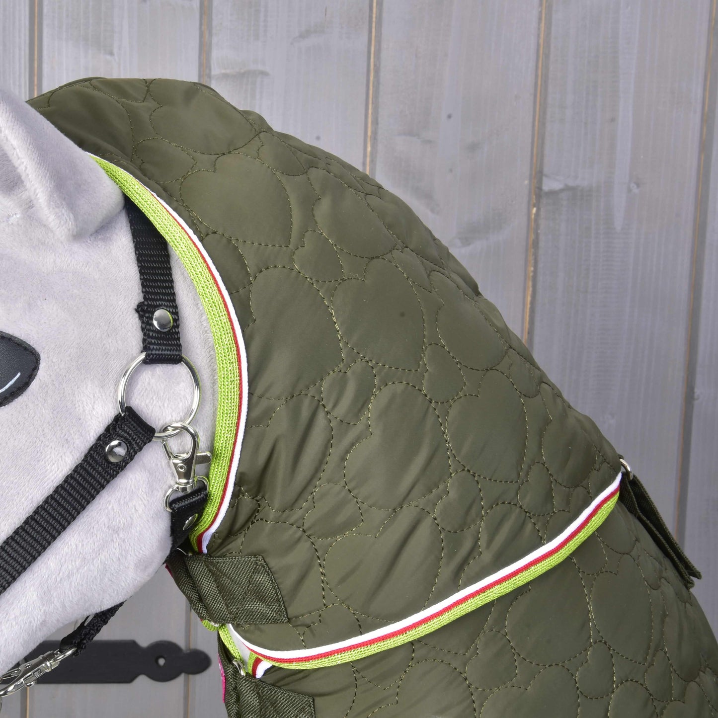 Detailný záber na psa s Dekou s odopínateľným krkom Rubby od Hobby Horse LarDen v olivovo zelenej farbe s neónovo zeleným zipsom a bielo-červeným lemom, stojaceho pred sivým dreveným pozadím.