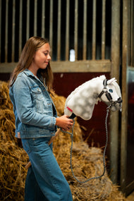 Dievča v džínsovej bunde stojí pred balíkmi slamy v stajni a drží Hobby Horse Elektra with Black Halter od Hobby Horse LarDen s bielou strakatou plyšovou hlavou, modrým okom a bielou priadzovou hrivou, s ohlávkou Graphite Full s kožušinou z koženky a pleteným vodítkom