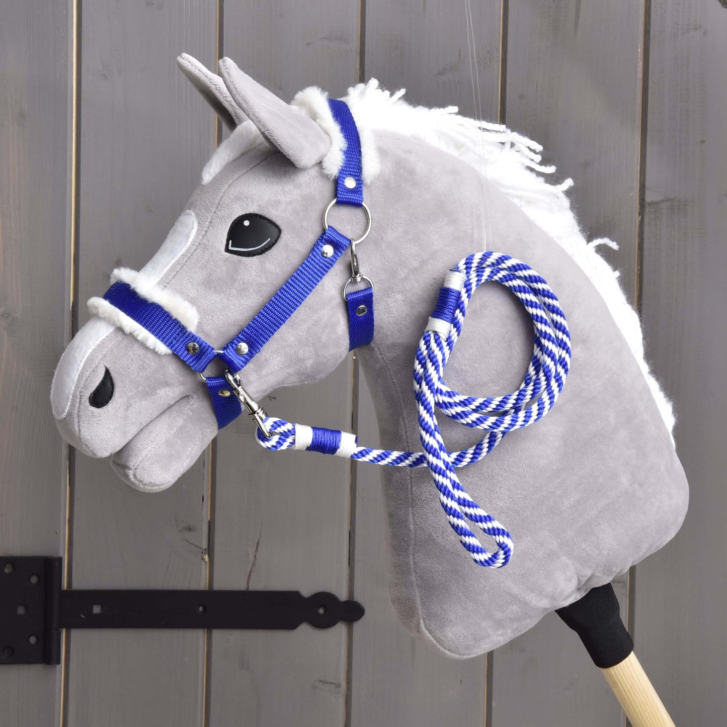 Set Essential Blue od Hobby Horse LarDen so sivým plyšovým Hobby Horse s bielou hrivou, modrou ohlávkou s kožušinovým lemom a modro-bielymi povrazovými opratami na drevenej tyči, zobrazený pred sivým dreveným plotom.