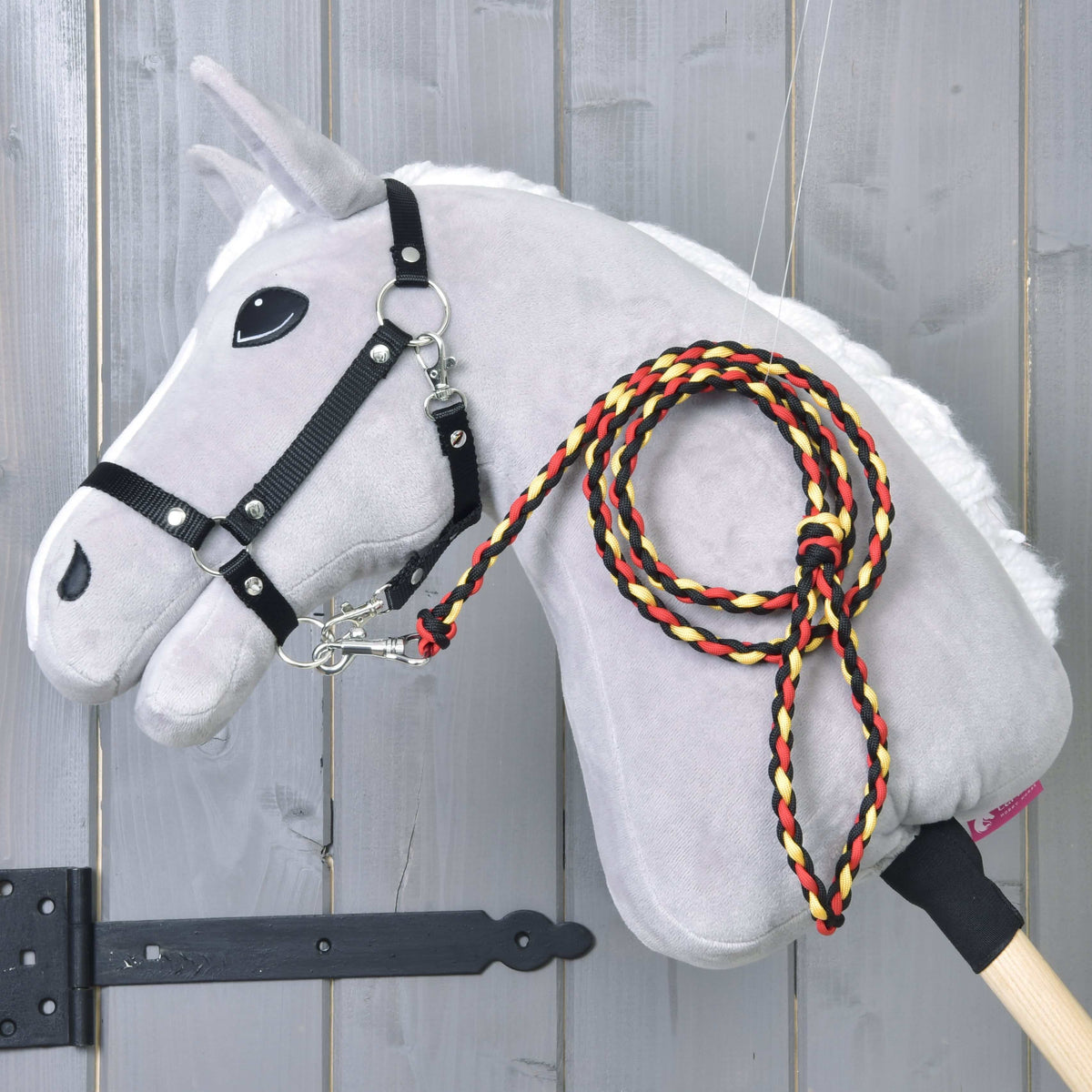 Hobby Horse LarDen "Hobby Horse - vodidlo Flag", sivý plyšový Hobby Horse s tyčovým telom, bielou hrivou, čiernou uzdečkou a pleteným červeným, žltým a čiernym vodidlom, zobrazený na drevenom pozadí.
