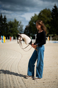Dievča v čiernom tričku stojí na pieskovej jazdiarni pod búrkovou oblohou a drží Hobby Horse Frederik LD with black halter od Hobby Horse LarDen, za ním sú farebné skokové tyče, s krémovou plyšovou hlavou a tmavohnedou priadzovou hrivou, s kompletnou hnedou uzdečkou s nánosníkom