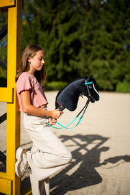 Dievča v ružovom tričku sa opiera o žltý skokový stojan a drží Hobby Horse Freya with Black Halter od Hobby Horse LarDen s čiernou plyšovou hlavou, bielou lysinou a čiernou priadzovou hrivou, s kompletnou tyrkysovou uzdečkou bez nánosníka