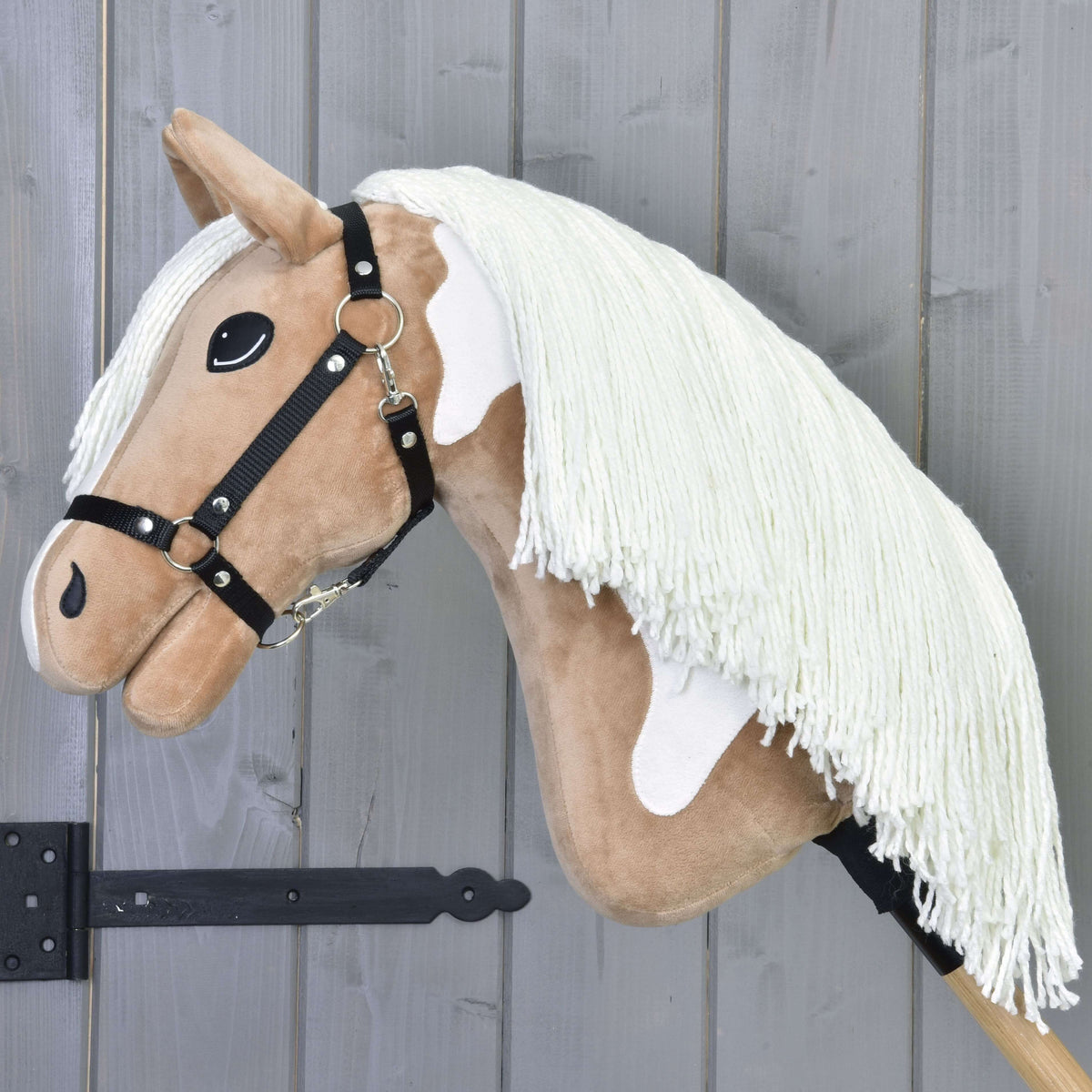 Hobby Horse Fury s čiernou ohlávkou od Hobby Horse LarDen má svetlohnedú tvár, biele škvrny, dlhú bielu hrivu a je nasadený na drevenej tyči, tu zobrazený pred sivou drevenou stenou.