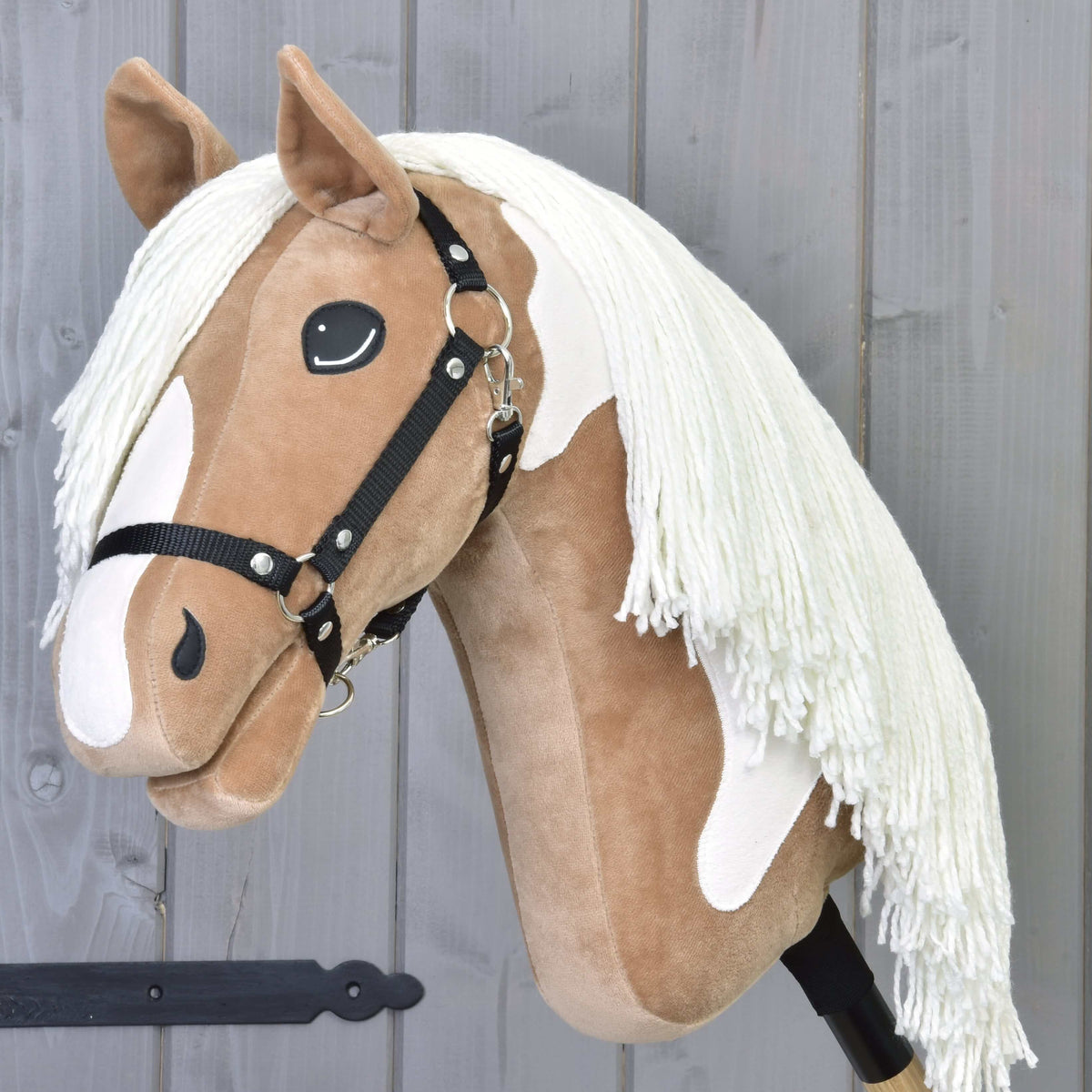 Hobby Horse Fury s čiernou ohlávkou od Hobby Horse LarDen má plyšovú svetlohnedú a bielu tvár, dlhú bielu hrivu, čiernu ohlávku a je nasadený na tyči, pripravený na vychádzky po chodníkoch pred sivým dreveným pozadím.