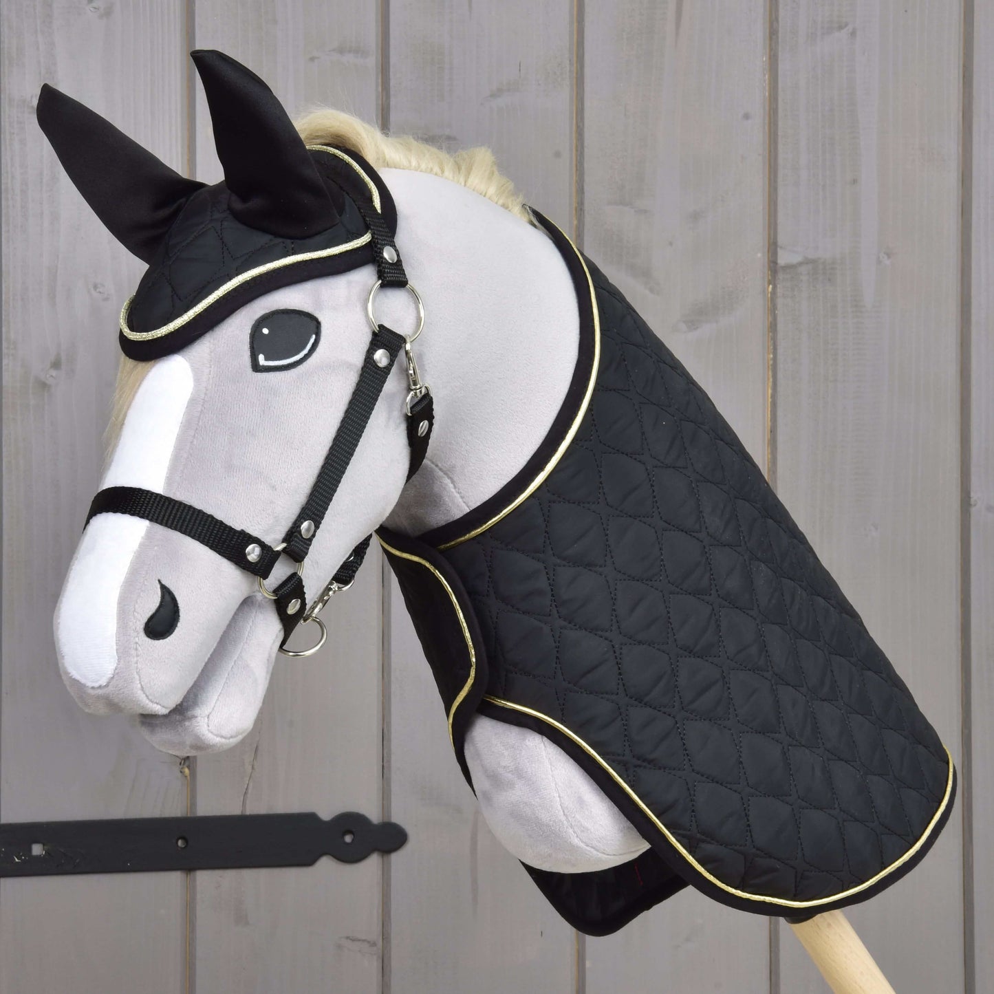 Hobby Horse LarDen Set Majestic obsahuje plyšového Hobby Horse s čiernym prešívaným poťahom, uzdečkou, opratami a krytmi uší proti hmyzu. Má biele a sivé znaky, svetlú hrivu a je zobrazený na drevenom pozadí.