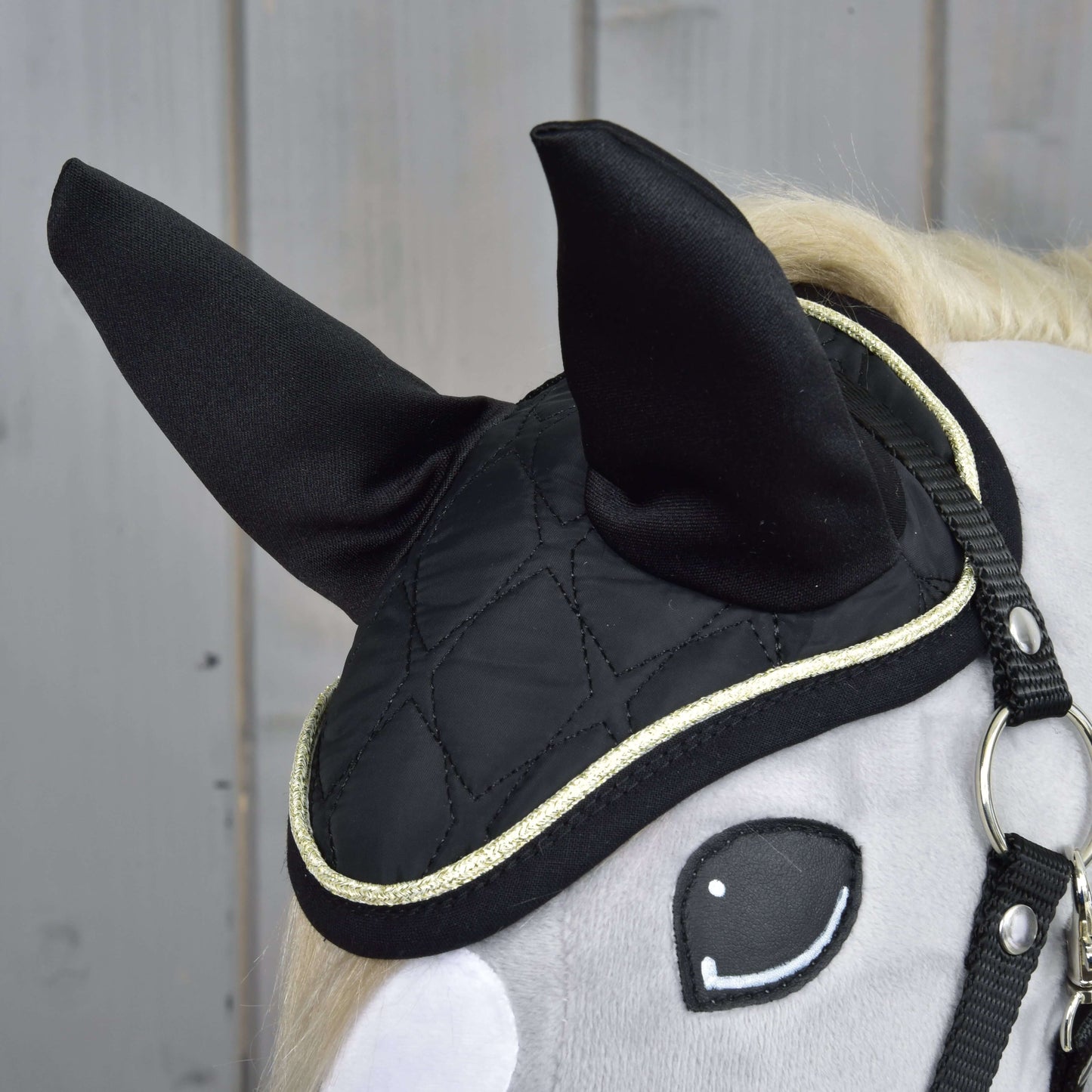 Detail hlavy sivého hračkárskeho koňa s čiernou prešívanou čabrakou Set Majestic od Hobby Horse LarDen so zlatým lemom, vztýčenými ušami a jednoduchou čiernou uzdečkou, zobrazený na drevenom pozadí.