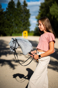 Dievča v ružovom tričku vedie Hobby Horse Grace with Black Halter od Hobby Horse LarDen cez pieskovú jazdiareň so sivou plyšovou hlavou a svetlosivou priadzovou hrivou, s kompletnou uzdečkou De Luxe s dvoma čelenkami
