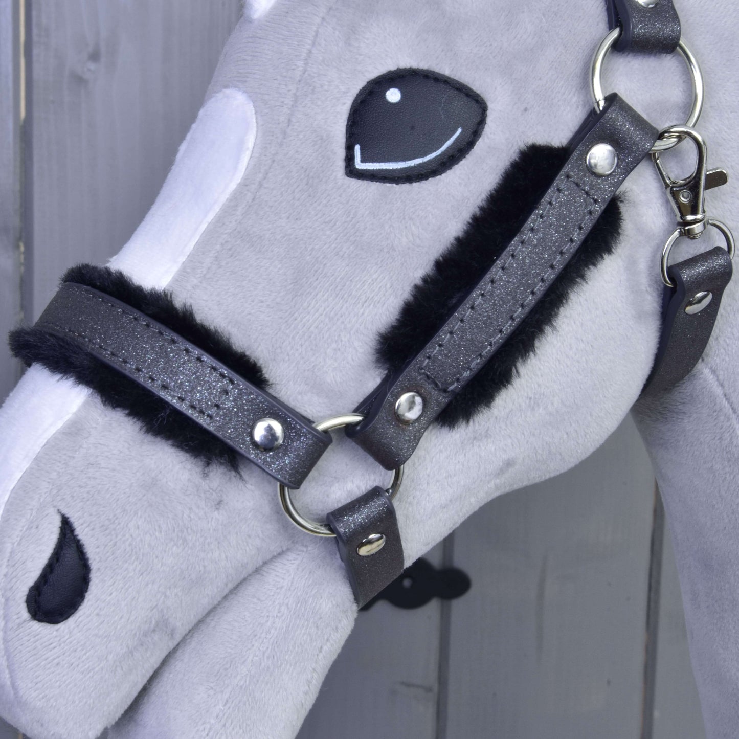 Detail sivobielej plyšovej hlavy Hobby Horse s Ohlávkou s barančekom z koženky Graphite Full od Hobby Horse LarDen a vyšívanými detailmi tváre na drevenom pozadí.
