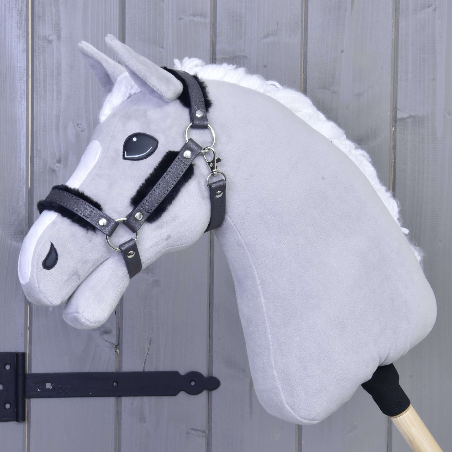 Ohlávka s barančekom z koženky Graphite Full od Hobby Horse LarDen na drevenej tyči, opretá o svetlosivú drevenú stenu, so sivou plyšovou hlavou s bielou hrivou, čiernou uzdečkou a ohlávkou z koženky.
