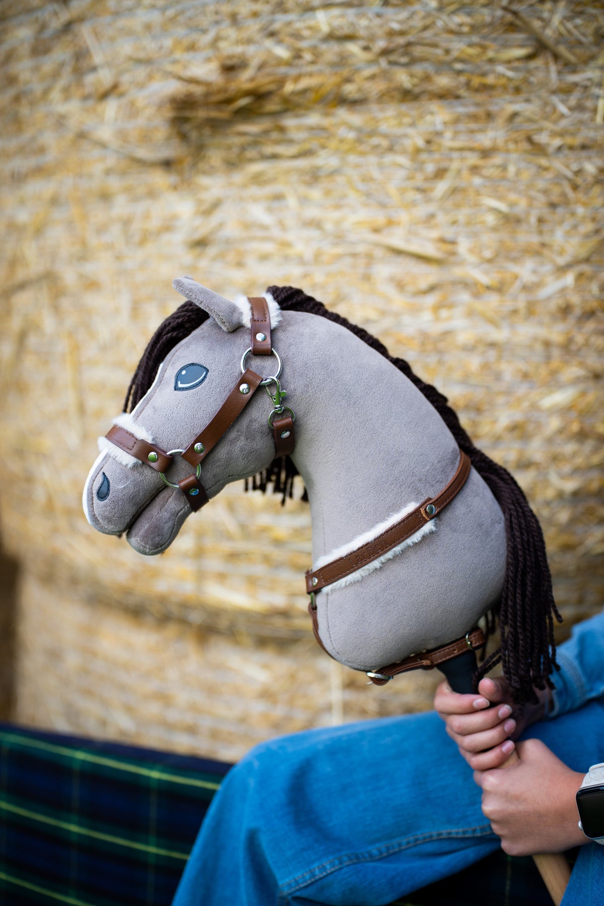 Hlava Hobby Horse Guccini od Hobby Horse LarDen pred balíkmi slamy so sivým plyšom, bielym pásikom na nose a tmavohnedou hrivou z priadze, s ohlávkou Walnut s kožušinou a náprsným remeňom Walnut s kožušinou
