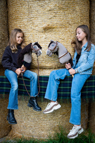 Dve dievčatá sedia na balíkoch slamy na tartanovej deke a každé drží Hobby Horse Guccini od Hobby Horse LarDen so sivou hlavou v štýle holštajnského koňa, bielym pásikom na nose a dlhou tmavohnedou hrivou z priadze, s ohlávkou Walnut s kožušinou, náprsným remeňom Walnut s kožušinou a vodiacim lanom Vivaldi