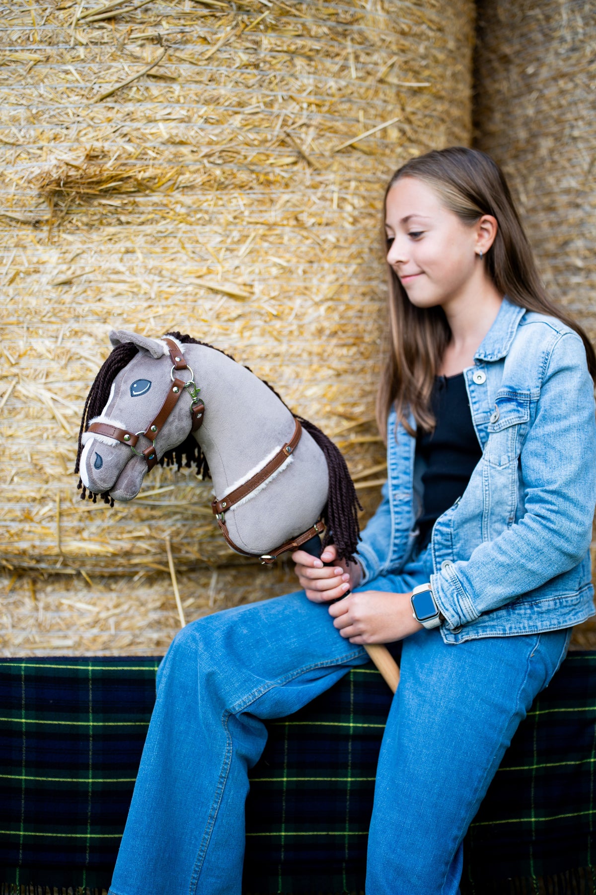 Dievča v rifľovej bunde sedí na balíkoch slamy a drží Hobby Horse Guccini od Hobby Horse LarDen so sivou hlavou, bielym pásikom na nose, dlhou tmavohnedou hrivou z priadze a drevenou tyčou, s ohlávkou Walnut s kožušinou a náprsným remeňom Walnut s kožušinou
