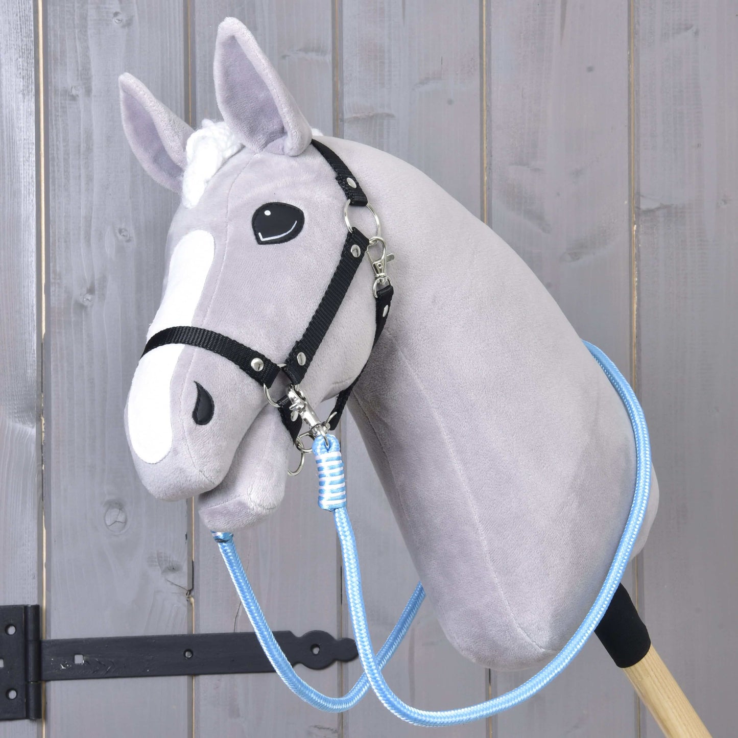 Opraty Heaven od Hobby Horse LarDen na sivom plyšovom Hobby Horse s bielym mulcom, čiernou uzdečkou a modrými polypropylénovými lanovými oprátami, na drevenej tyči pred dreveným obložením.