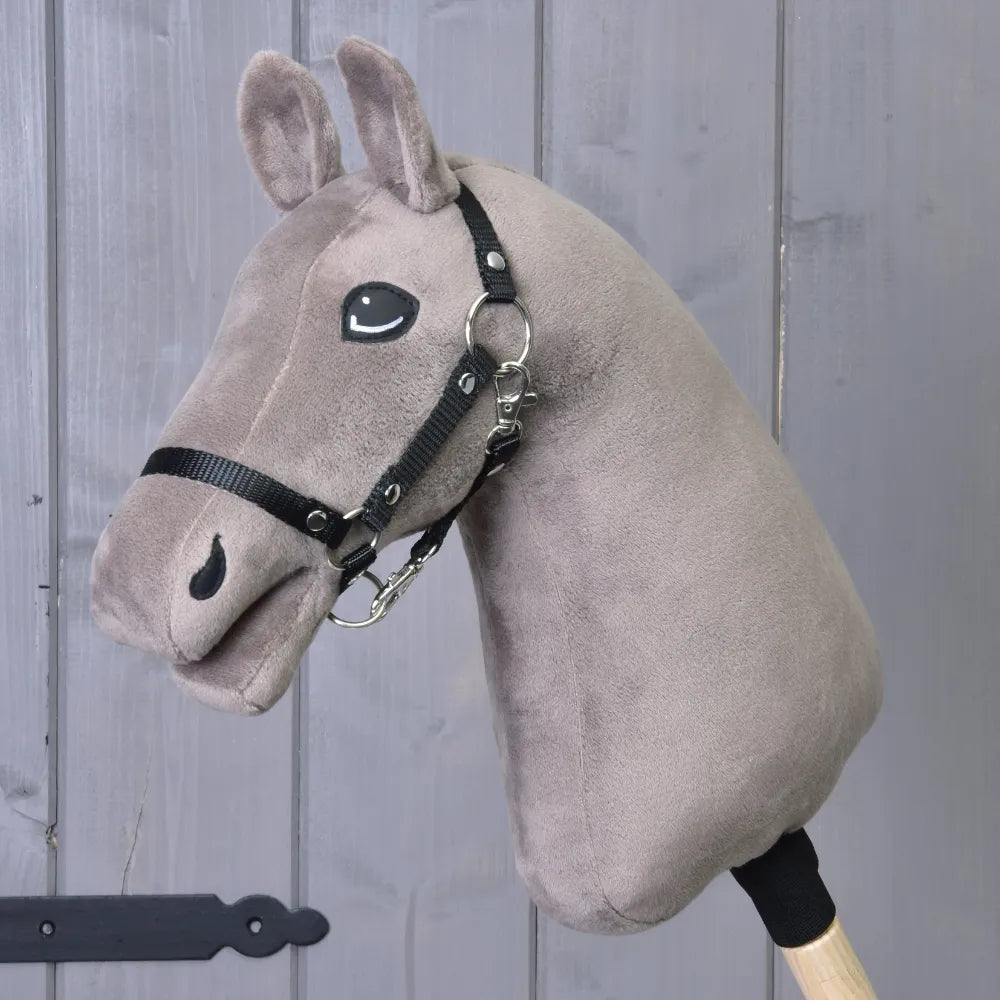 The Build your horse by related_to_12441055625556 features a plush taupe hobby horse head with a black yarn mane and bridle on a wooden stick, shown in front of a gray wooden wall.