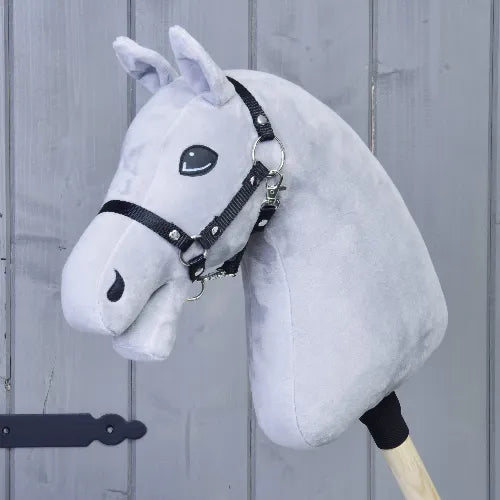 The Build your horse by related_to_12441055625556 is a light grey custom hobby horse with a black bridle and beige yarn mane on a wooden stick, shown in front of a gray wooden wall.