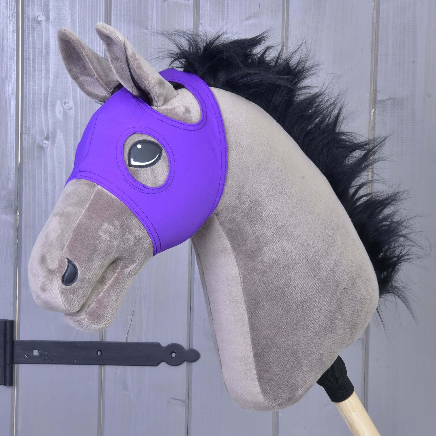 Plyšový Hobby Horse s čiernou hrivou, sivou hlavou a Maskou bez uší Indigo od Hobby Horse LarDen v indigovej farbe, vyrobenou z prateľného materiálu PES, nasadený na drevenej tyči na pozadí sivej drevenej steny.