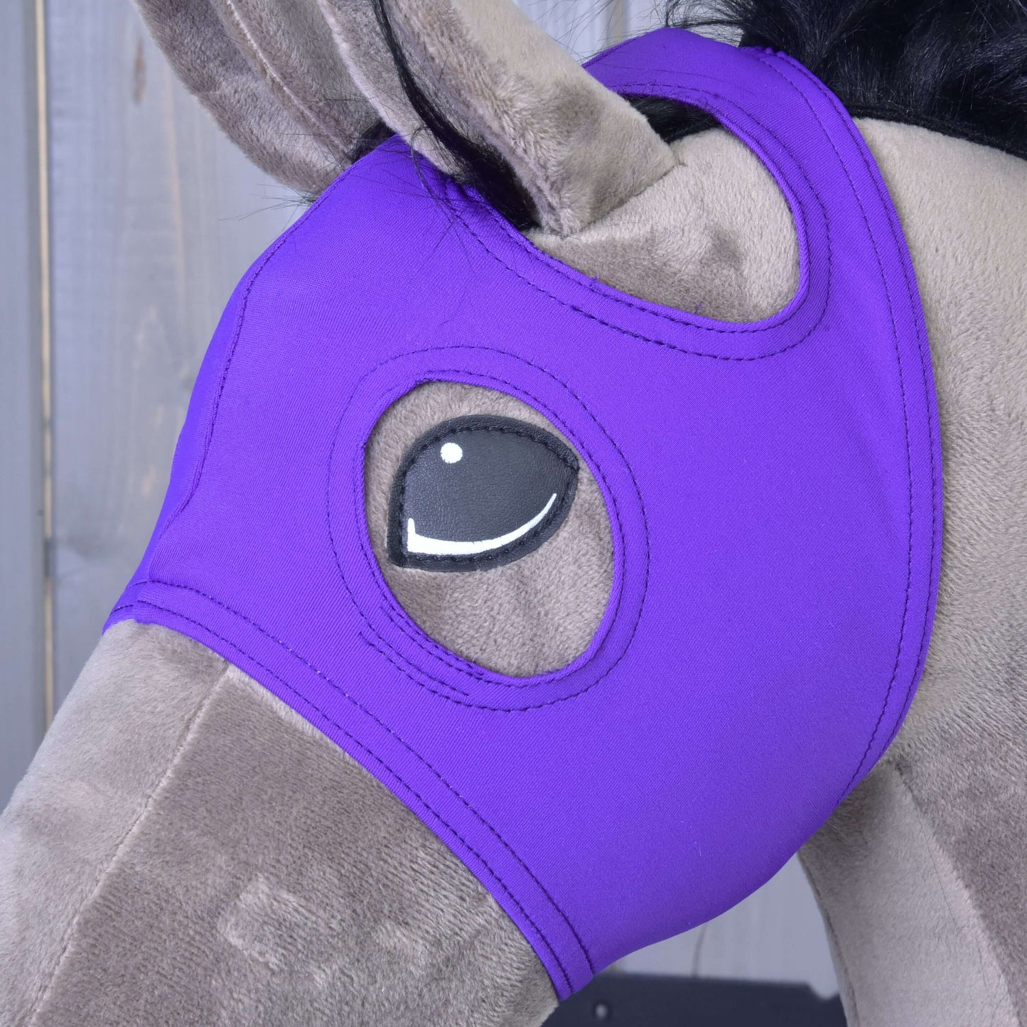 Plyšový hračkársky kôň má nasadenú Masku bez uší Indigo od Hobby Horse LarDen, vyrobenú z prateľného PES, zakrývajúcu hlavu s otvormi pre oči, na drevenom pozadí.