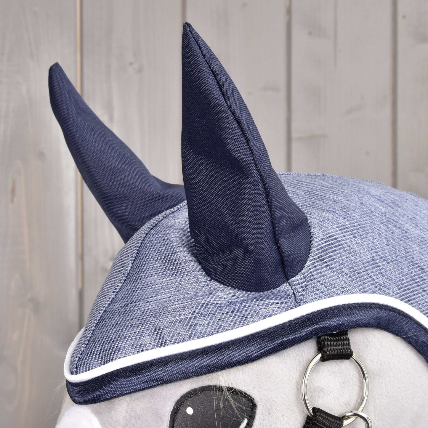 Detail šedo-tmavě modré čepice Hobby Horse LarDen Raincoat Jeans se dvěma špičatými ušima, bílým lemováním a kovovým kroužkem – ideální ke kombinaci s celoroční pláštěnkou vašeho Hobby Horse – zobrazeno na dřevěném pozadí.