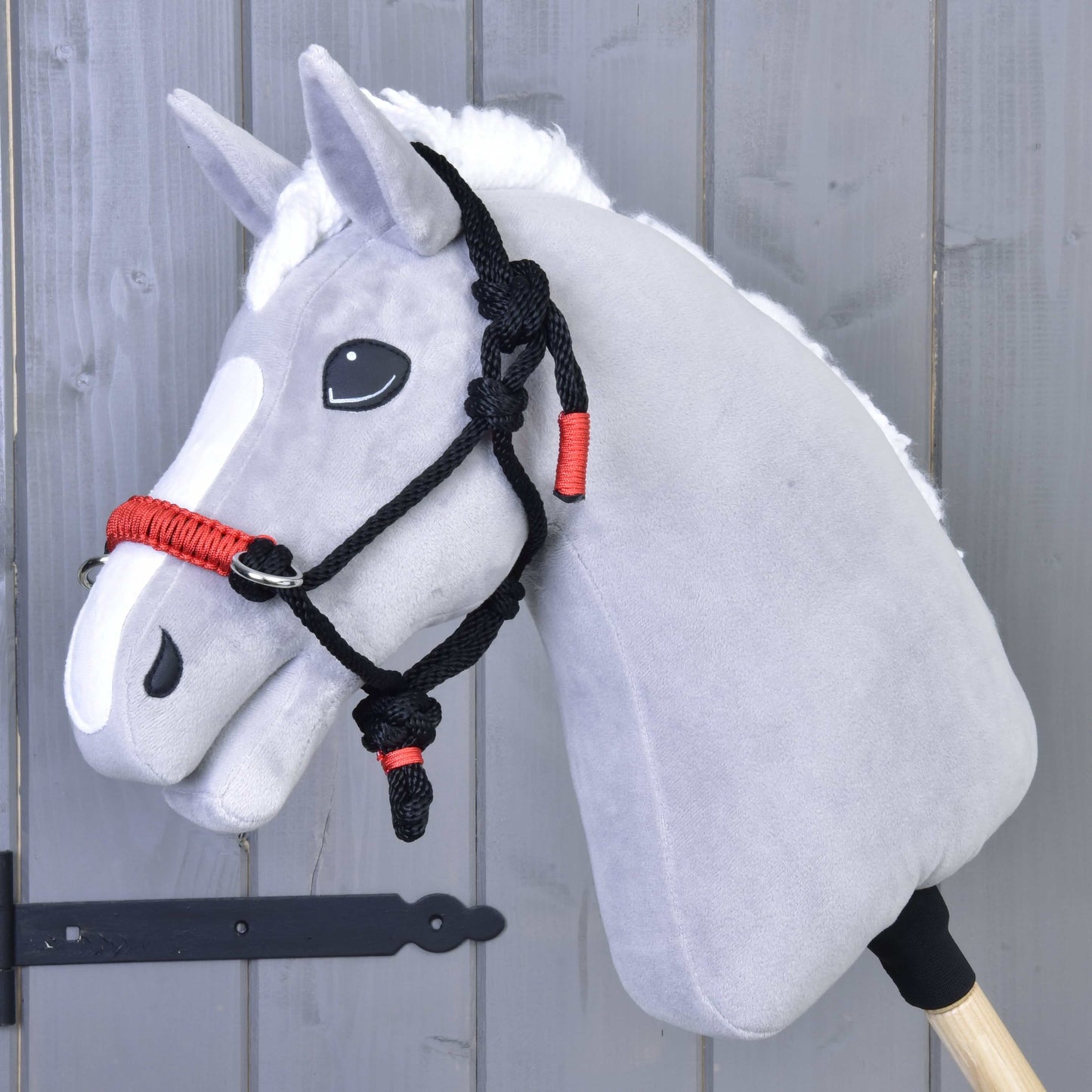 Hobby Horse LarDen Parelka Jiskra na sivom plyšovom Hobby Horse s bielou hrivou a čierno-červenou parelkou, nasadenom na drevenej tyči pred sivou drevenou stenou.