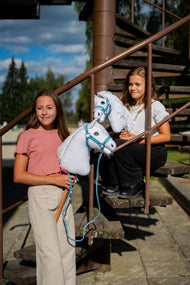 Dve dievčatá s bielymi Hobby Horses od Hobby Horse LarDen na hrdzavom kovovom schodisku, vpredu Hobby Horse Lady with Black Halter s bielou plyšovou hlavou a bielou priadzovou hrivou, s ohlávkou Pastell Blue s kožušinou z koženky a pleteným vodítkom Double Blue