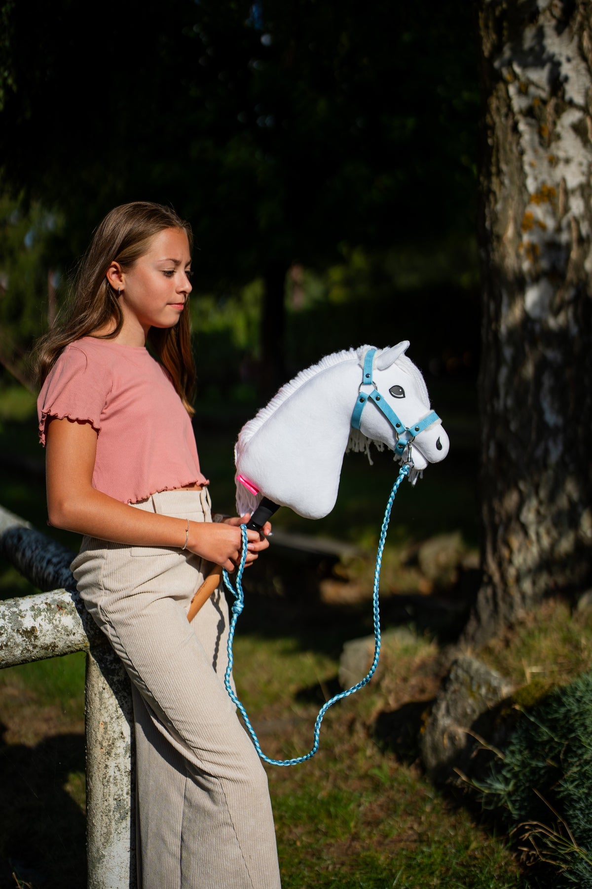 Dievča v ružovom tričku stojí pri machom obrastenom strome a drží Hobby Horse Lady with Black Halter od Hobby Horse LarDen, vodítko visí v slučke, s bielou plyšovou hlavou a bielou priadzovou hrivou, s ohlávkou Pastell Blue s kožušinou z koženky a pleteným vodítkom Double Blue