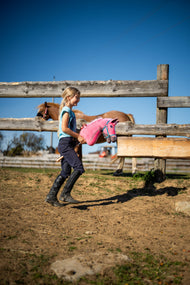 Lifestyle photo: a girl in riding clothes walks across a sandy paddock with the Hobby Horse LarDen Pinkie with a glittery halter by Hobby Horse LarDen - pink plush head with a pink and white yarn mane - shown here with the Halter turquoise.
