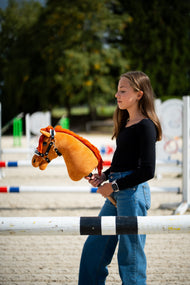 Dievča v čiernom tričku stojí za skokovou tyčou na pieskovej jazdiarni a drží Hobby Horse Little Angel with Black Halter od Hobby Horse LarDen s gaštanovohnedou plyšovou hlavou a jasne červenou priadzovou hrivou, s ohlávkou Espresso s kožušinou z koženky