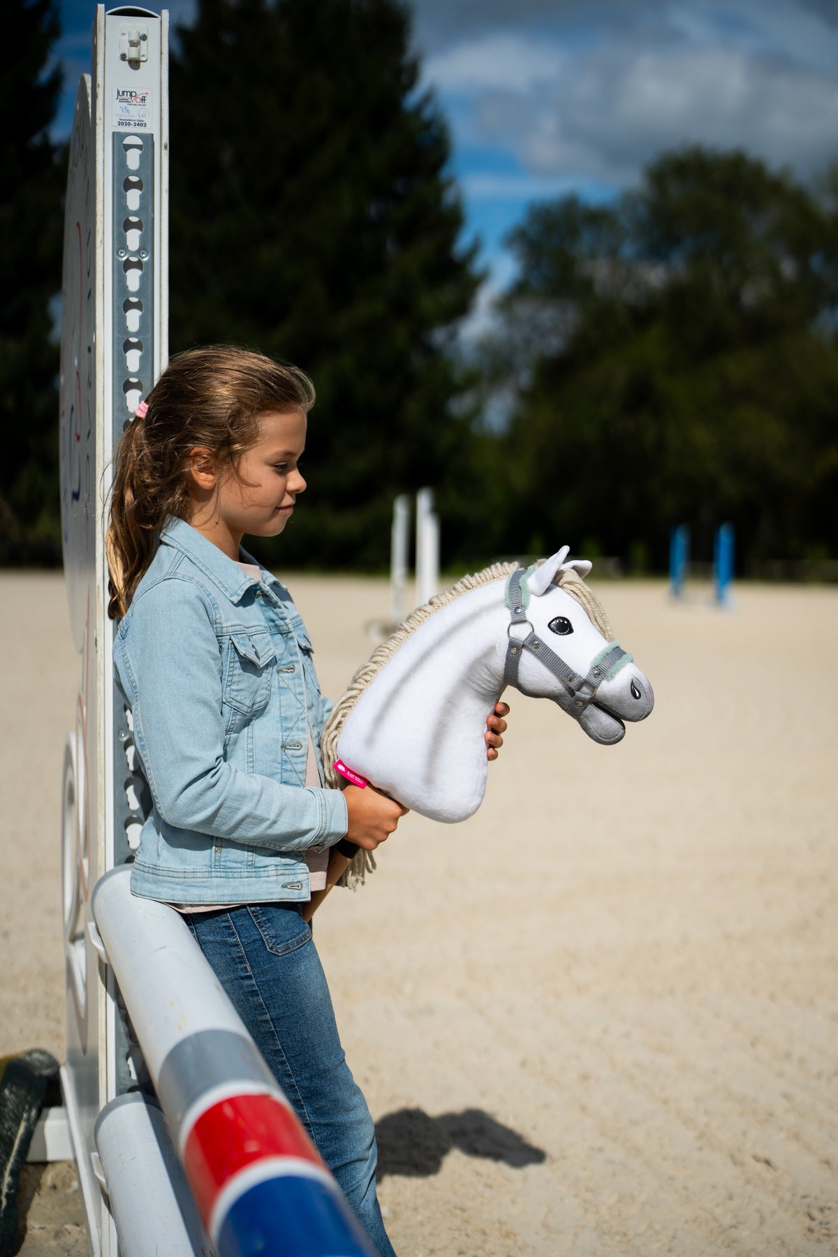 Dievča v džínsovej bunde stojí pri skokovom stojane na pieskovej jazdiarni a drží Hobby Horse Little Lady with Black Halter od Hobby Horse LarDen s bielou plyšovou hlavou a krémovou priadzovou hrivou, so sivou ohlávkou s mätovou kožušinou