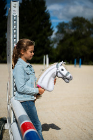 Dievča v džínsovej bunde stojí pri skokovom stojane na pieskovej jazdiarni a drží Hobby Horse Little Lady with Black Halter od Hobby Horse LarDen s bielou plyšovou hlavou a krémovou priadzovou hrivou, so sivou ohlávkou s mätovou kožušinou