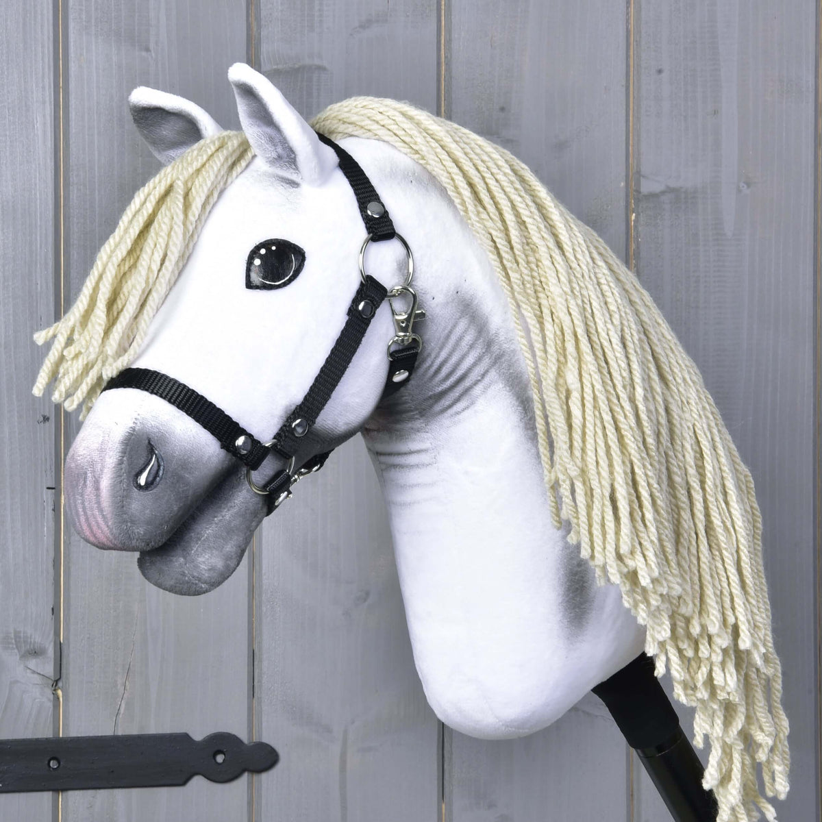 Hobby Horse Little Lady s čiernou ohlávkou od Hobby Horse LarDen má biele telo a dlhú krémovo sfarbenú hrivu z priadze, stojí pred drevenými panelmi a odráža eleganciu drezúry Pura Raza Española.