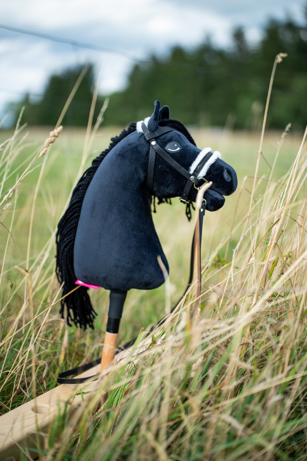 Hobby Horse Mahal with Black Halter od Hobby Horse LarDen stojí vo vysokej suchej tráve na lúke s čiernou plyšovou hlavou, čiernou priadzovou hrivou a drevenou tyčou, s kompletnou uzdečkou s kožušinou