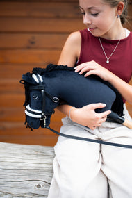 Dievča v bordovom tričku drží Hobby Horse Mahal with Black Halter od Hobby Horse LarDen v náručí pred drevenou stenou s čiernou plyšovou hlavou, čiernou priadzovou hrivou a bielym kožušinovým nánosníkom, s kompletnou uzdečkou s kožušinou