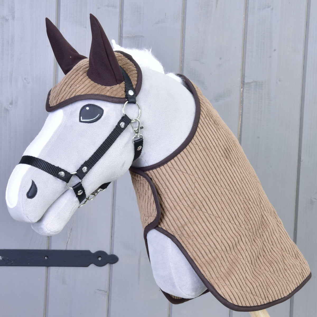 Set Manchester Le Chocolate od Hobby Horse LarDen s plyšovým Hobby Horse s bielou tvárou, čiernym postrojom, hnedými ušami a štruktúrovanou hnedou látkou na hlave a krku, doplnený Hobby Horse dekou, na drevenej tyči pred sivou drevenou stenou.