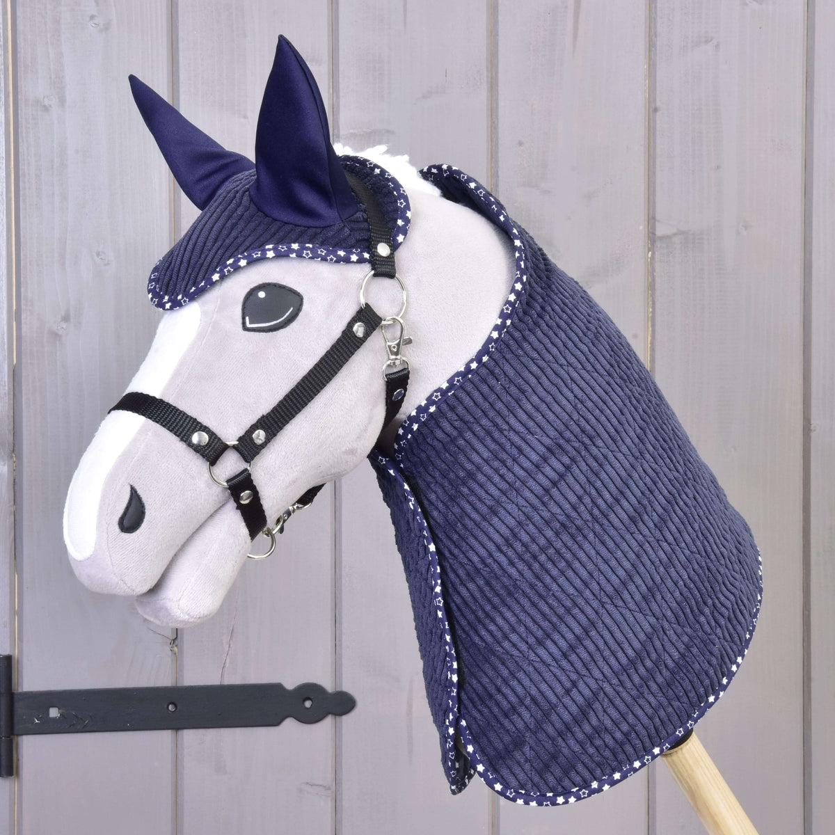 Hobby Horse LarDen Set Manchester Blue Star obsahuje sivého Hobby Horse s čiernou uzdečkou a opratami, tmavomodrú prešívanú deku a čabraku s bielym lemom, zobrazené na drevenej tyči pred svetlým dreveným obložením.