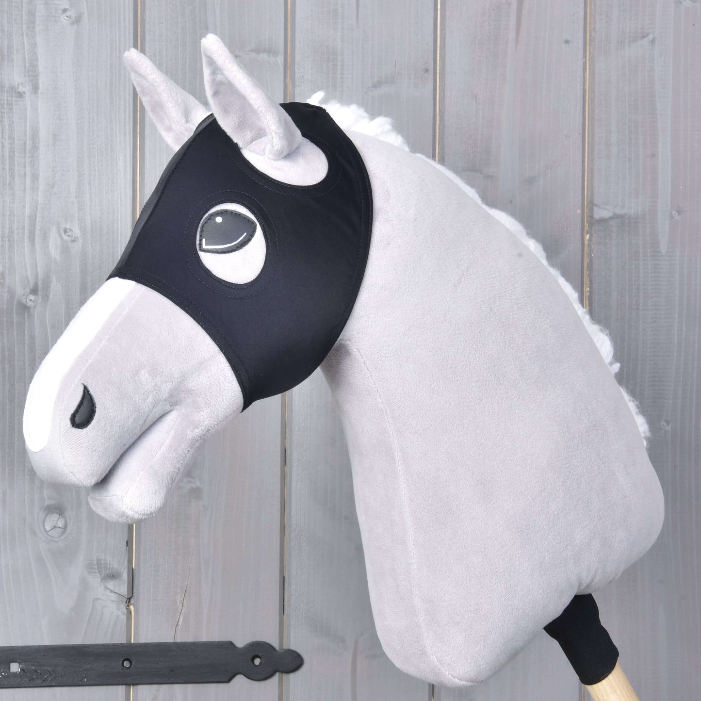 Plyšový Hobby Horse so svetlosivou srsťou, bielou hrivou a čiernou uzdečkou má na sebe masku Maska bez uší Flamingo od Hobby Horse LarDen a je nasadený na drevenej tyči pred pozadím zo sivých drevených panelov.