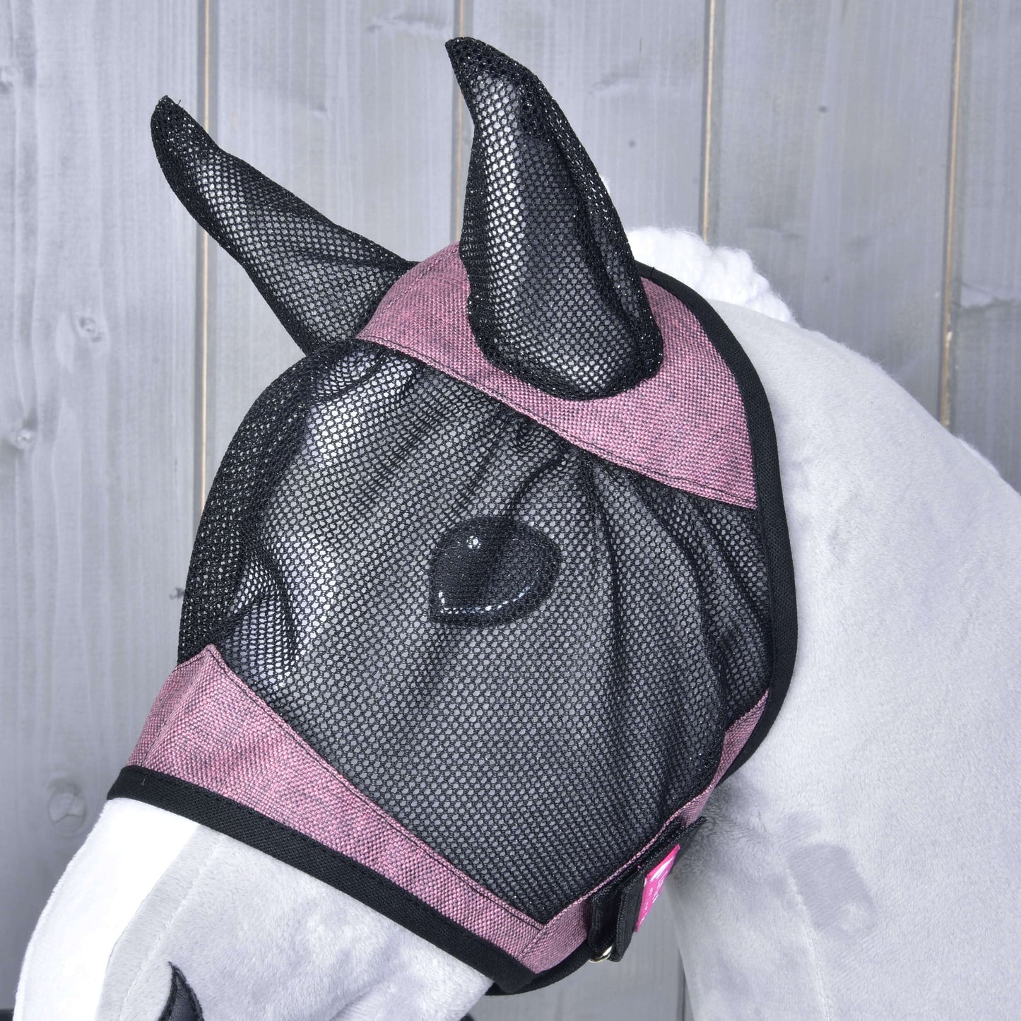Biely kôň má na sebe Maska proti hmyzu de Luxe Pink od Hobby Horse LarDen, prateľnú čierno-ružovú sieťovanú masku zakrývajúcu uši a tvár, na svetlosivom drevenom pozadí.