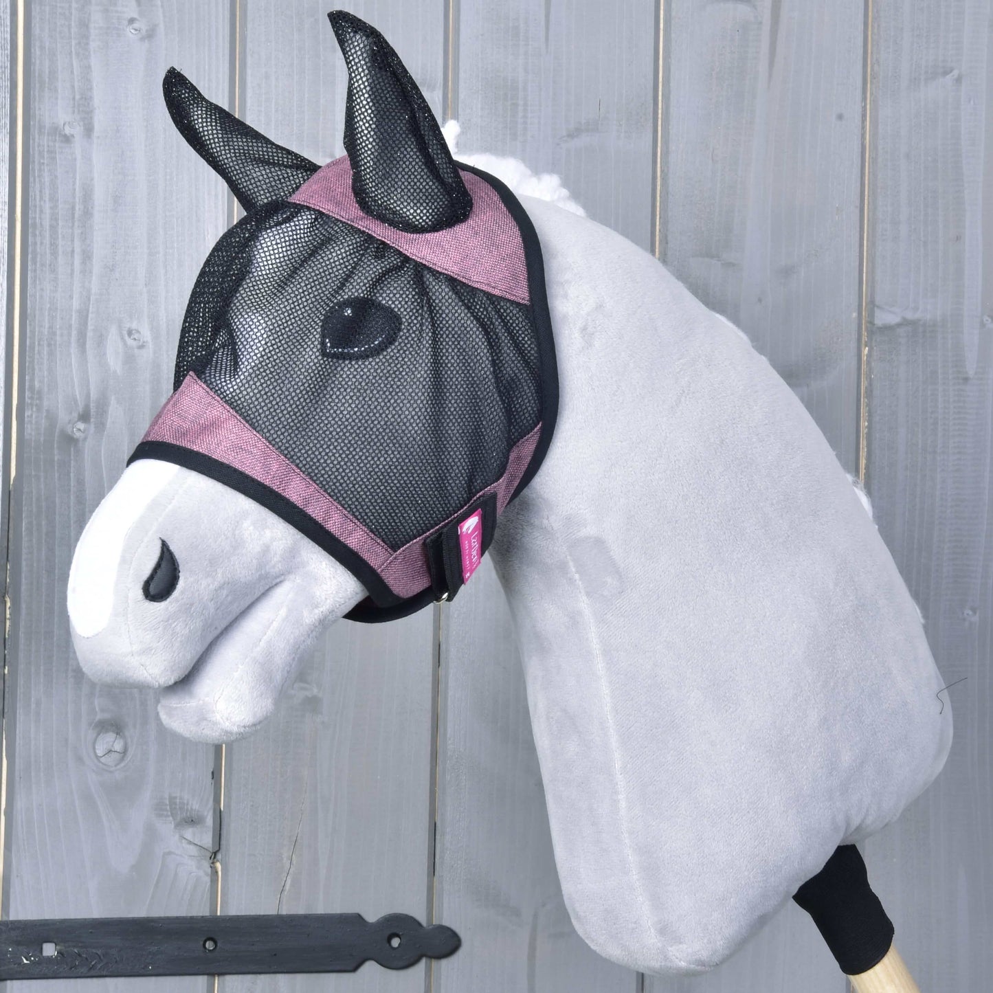 Maska proti hmyzu de Luxe Pink od Hobby Horse LarDen na sivom plyšovom Hobby Horse s ružovým lemovaním a prateľnou maskou proti hmyzu, nasadenom na drevenej tyči, na pozadí z drevených dosiek.
