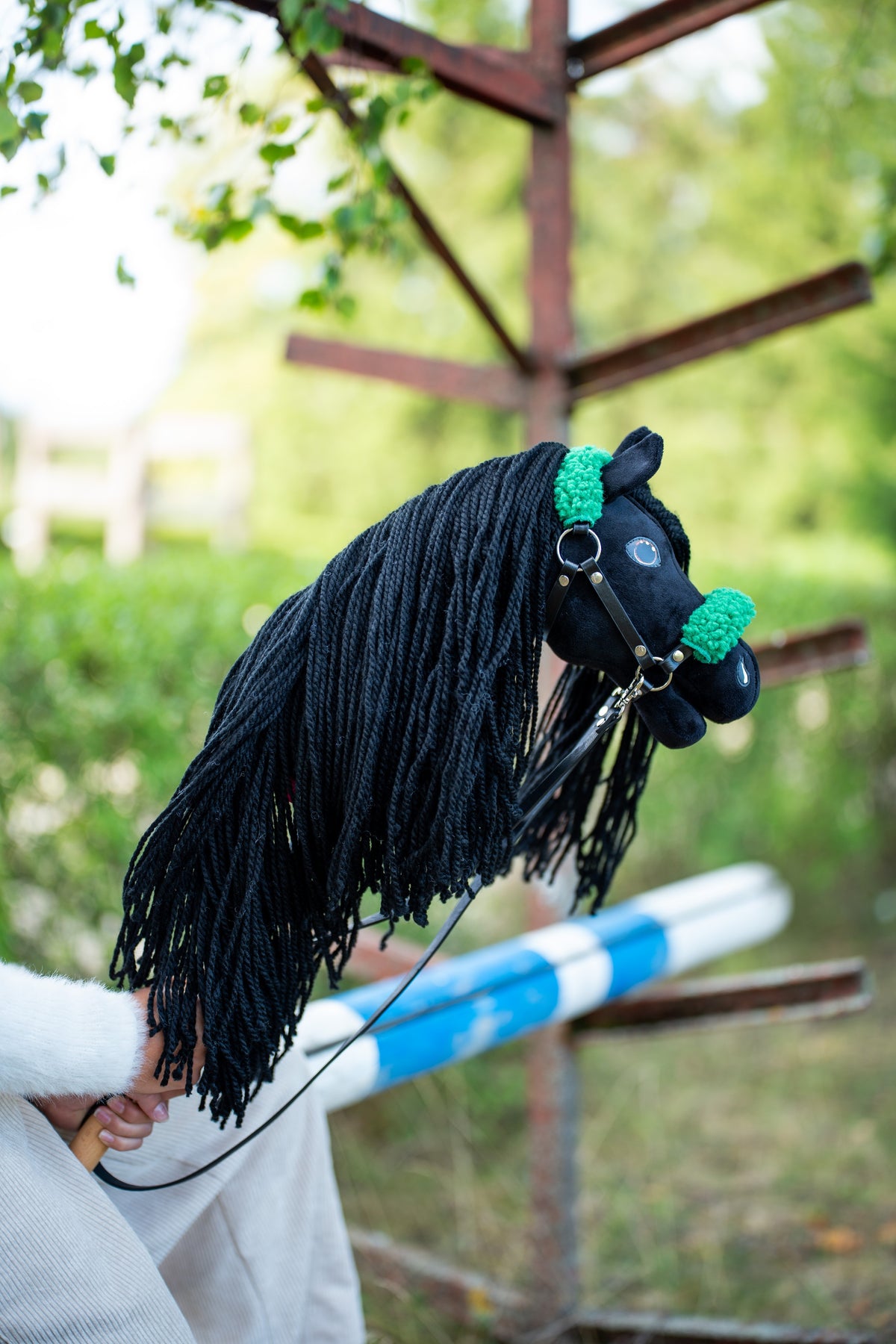 Hlava a dlhá hriva Hobby Horse Maximus with Black Halter od LarDen zdvihnuté pri drevenej prekážke, v pozadí zeleň – čierny plyš a čierna priadzová hriva – s čiernou koženkovou ohlávkou so zelenou kožušinou a čiernymi koženkovými oťažami