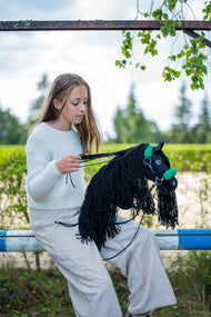 Dievča vo svetlom svetri sedí na ohradníku a zhŕňa opraty Hobby Horse Maximus with Black Halter od Hobby Horse LarDen s čiernou plyšovou hlavou a dlhou čiernou priadzovou hrivou, s čiernou koženkovou ohlávkou so zelenou kožušinou a čiernymi koženkovými opratami