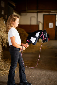 Dievča v bielom tričku vedie Hobby Horse Merlin s Black Halter od Hobby Horse LarDen popri balíkoch slamy v stajni – čiernobiela strakatá plyšová hlava, čierna priadzová hriva – s ružovou súpravou LarDen Exclusive Pink