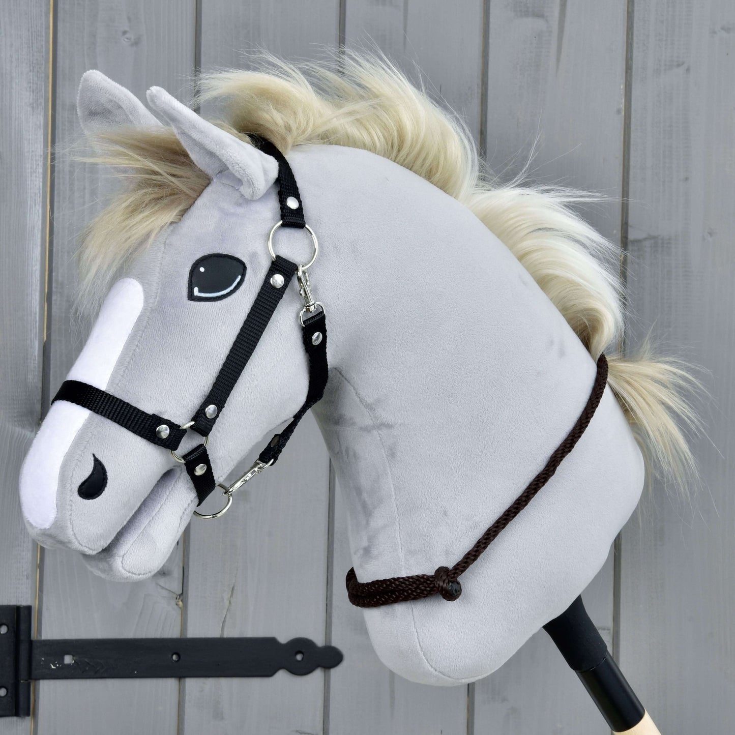 Nákrčník Chocolate od Hobby Horse LarDen so sivým plyšovým Hobby Horse, čiernou uzdečkou a krémovou povrazovou hrivou na drevenej tyči, na pozadí sivého dreveného panelu.