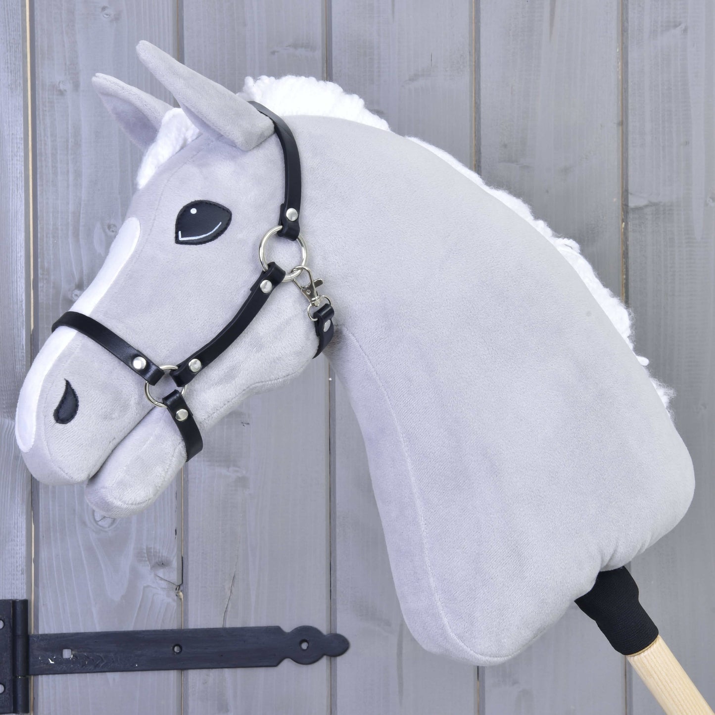 Sivý plyšový Hobby Horse od Hobby Horse LarDen s bielou hrivou a ohlávkou Ohlávka - čierna koženka, nasadený na drevenej tyči na pozadí drevených panelov.