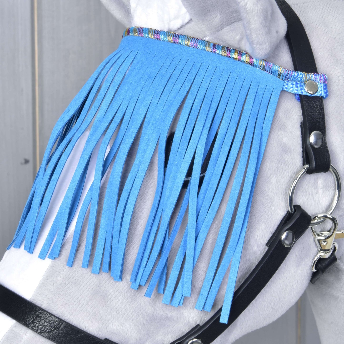 Detailní záběr koně s Ohlávkou černou s modrými třásněmi od Hobby Horse LarDen, s duhově lemovanou muší třásní na ochranu očí před mouchami, na pozadí světlé dřevěné stěny.