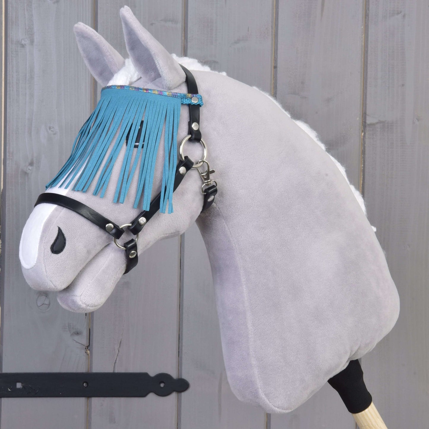 Šedý Hobby Horse LarDen s černou ohlávkou s tyrkysovými třásněmi stojí před dřevěnou stěnou.