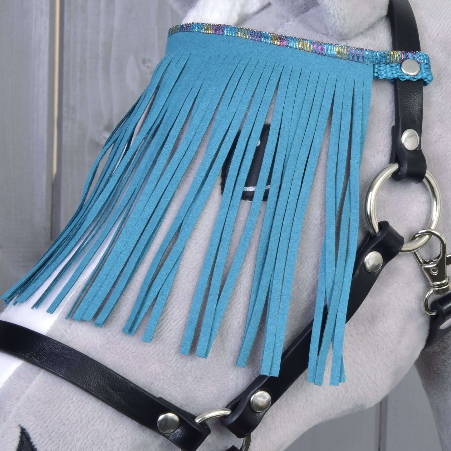 Detail koně s ohlávkou Hobby Horse LarDen, černou s tyrkysovými třásněmi, vedle pozadí z dřevěné stěny.