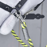 Detail kovové karabiny připevňující černý popruh ke žlutým, černým a šedým otěžím Hobby Horse - otěže proužek lemon od Hobby Horse LarDen. Pozadí tvoří rozmazané šedé dřevo a je vidět část měkkého bílého předmětu.