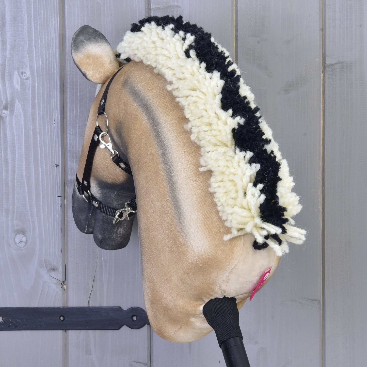 Hobby Horse Ozzy s čiernou ohlávkou od Hobby Horse LarDen s ručne vyrobenou béžovou tvárou, čiernou ohlávkou a huňatou čierno-bielou hrivou z priadze, nasadený na drevenej tyči pred stenou zo sivých drevených panelov.