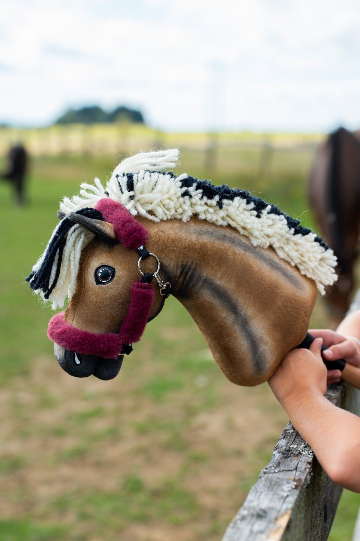 Hlava Hobby Horse Ozzy s Black Halter od Hobby Horse LarDen opretá o drevenú ohradu výbehu, za ňou usmiate dievča a pasúce sa kone – hnedý plyš s tmavým tieňovaním, krémová a čierna priadzová hriva – s halterom Teddy Maroon podšitým kožušinou